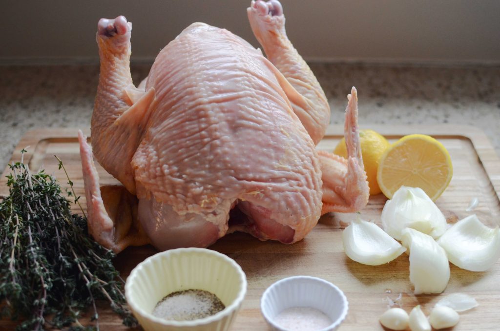 Whole raw chicken, cut lemon, thyme sprigs, onion, garlic cloves and small dishes with salt and black pepper