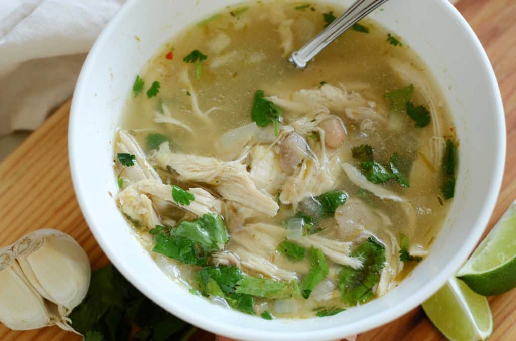 White chicken chili in white bowl with lime wedges and garlic cloves on wooden board
