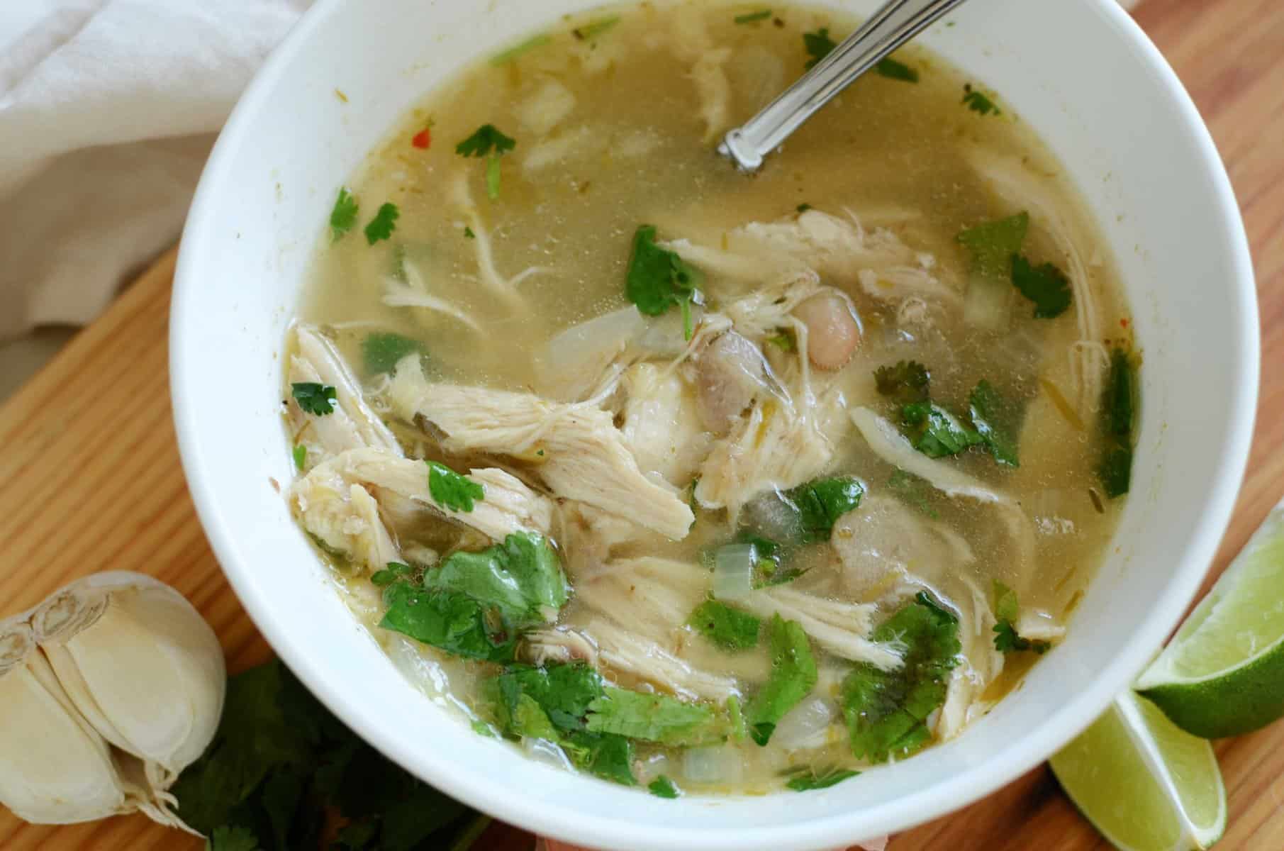 White chicken chili in white bowl with lime wedges and garlic cloves on wooden board