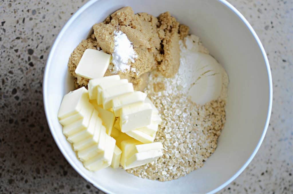 Flour, oatmeal, brown sugar, baking soda and sliced butter in white mixing bowl.