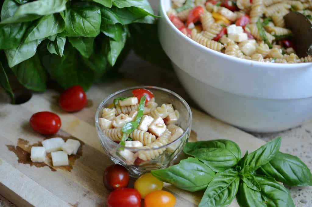 Caprese pasta salad in small glass dish with small tomatoes, fresh basil sprigs and diced fresh mozzarella all around. Large white bowl of pasta salad in background