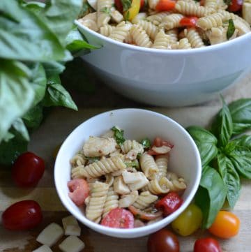 Caprese pasta salad in white bowl with tomatoes and basil sprigs scattered around bowl.