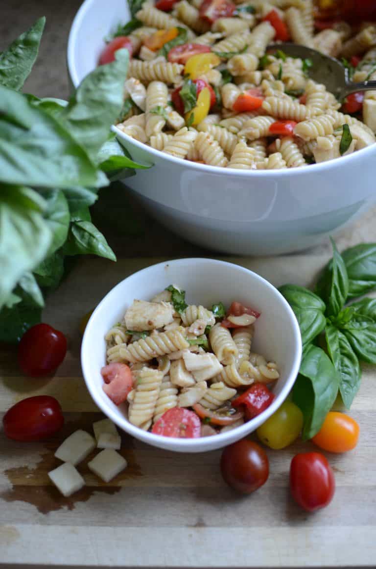 Caprese pasta salad in white bowl with tomatoes and basil sprigs scattered around bowl.