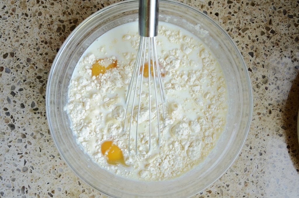 Milk, eggs, flour and sugar in glass mixing bowl with whisk.