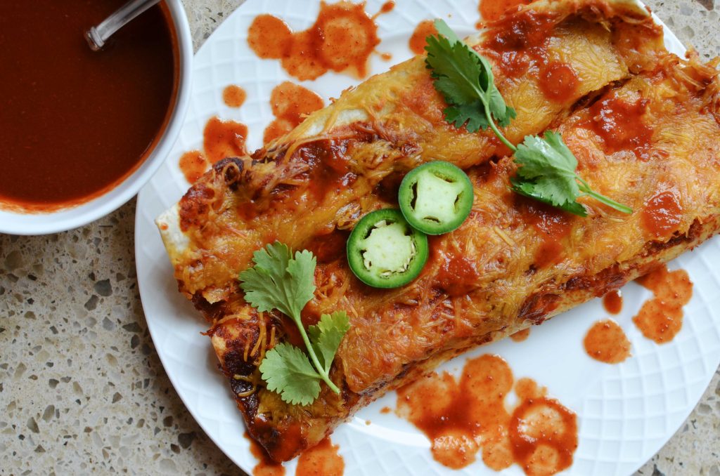 Two flour tortilla enchiladas with cilantro and sliced jalapeno peppers on white plate, with small white dish of enchilada sauce on the side.