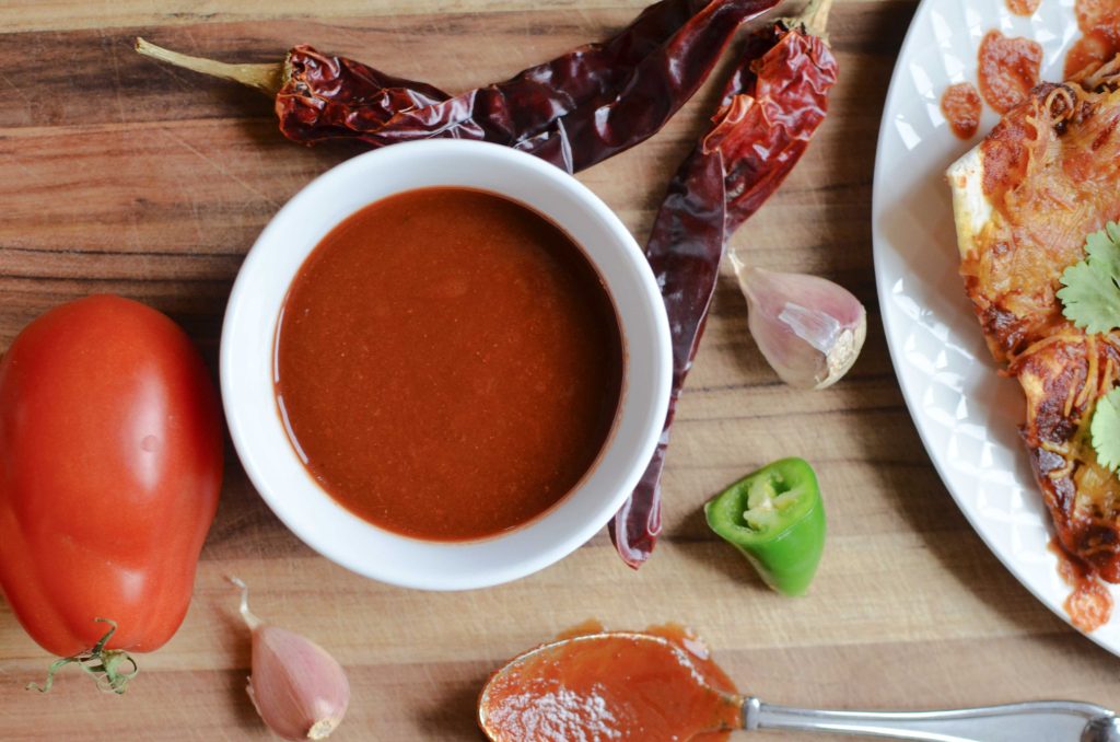 enchilada sauce in white bowl, with dried chilies, garlic cloves, tomato and spoon covered with enchilada sauce