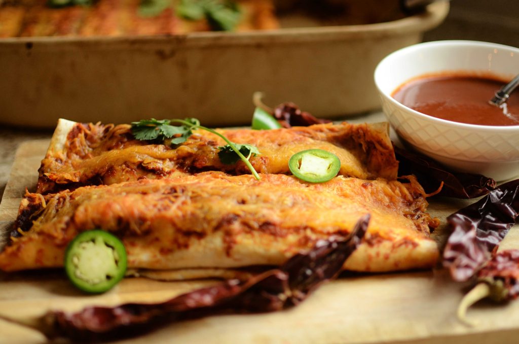 Enchiladas on wooden cutting board with cilantro and sliced jalapenos on top. Dried pepper in front of the enchilada
