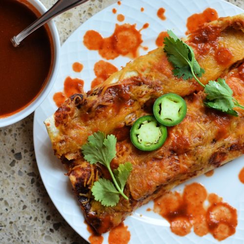 Enchiladas on plate with jalapeno slices and cilantro and a small bowl of enchilada sauce to the side.