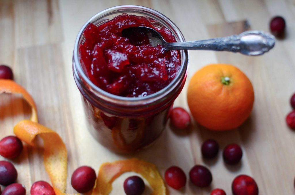 Half pint mason jar of cranberry sauce with spoon in jar. Orange, orange peel and cranberries all around jar. 