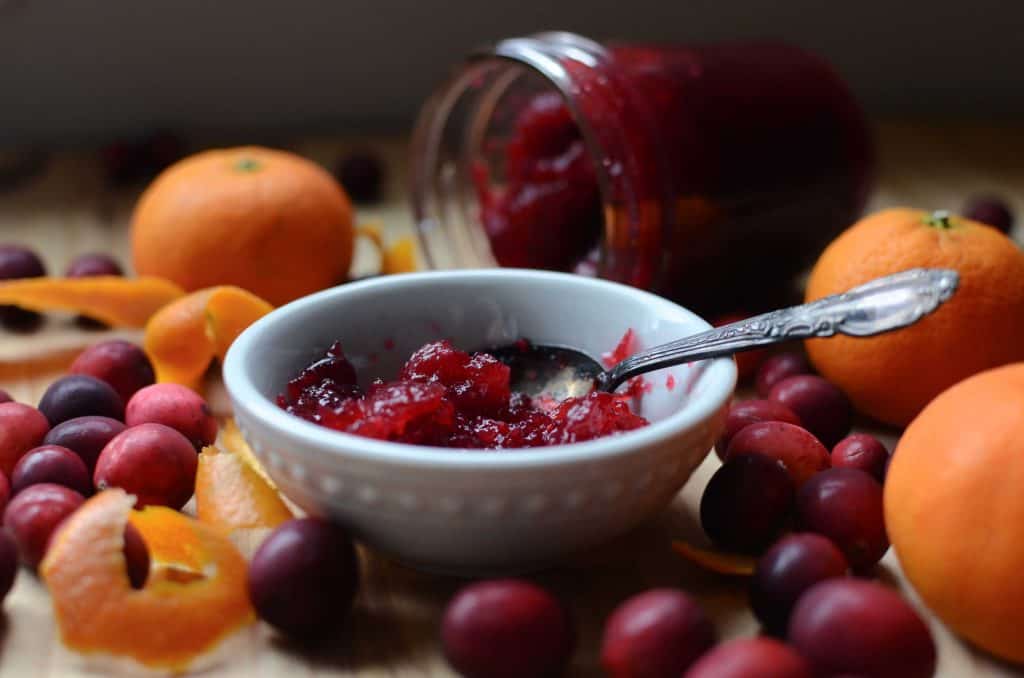 Small white dish with cranberry sauce. Jar with sauce behind and cranberries, oranges and orange rind around dish