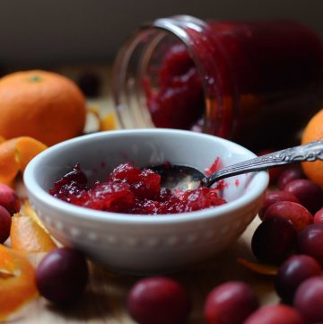 Small white dish with cranberry sauce. Jar with sauce behind and cranberries, oranges and orange rind around dish