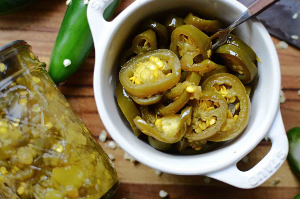 a small dish full of  candied jalapenos with a small fork, a jar of candied jalapenos and a jalapeno pepper