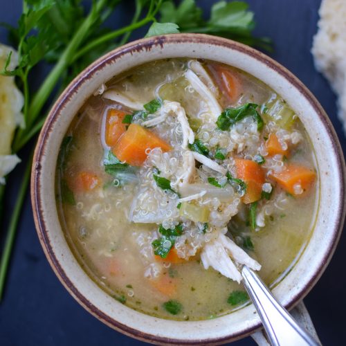 a cup of chicken quinoa soup with spoon, parley sprigs and half of a lemon