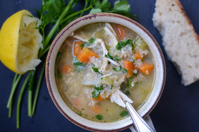 a cup of chicken quinoa soup with spoon, parley sprigs and half of a lemon