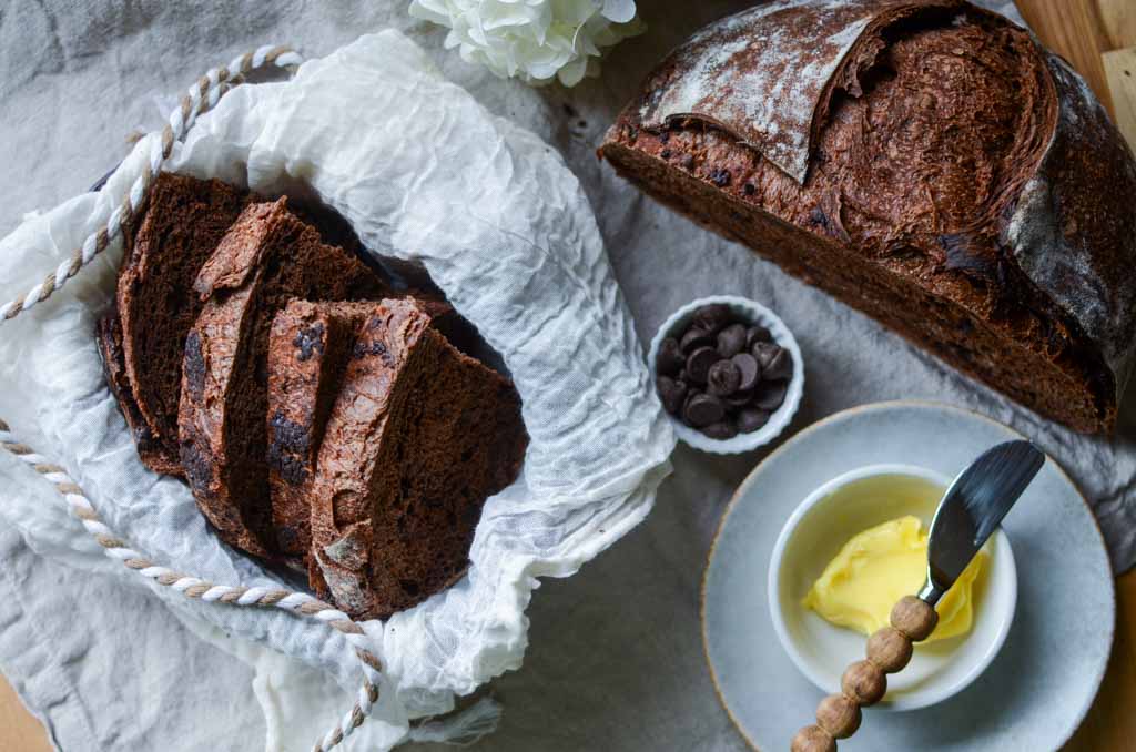 lined basket of sourdough chocolate bread slices and half loaf, bowl of chocolate chips, and a dish of butter and butter knife. 