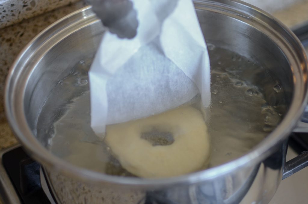 dropping a sourdough bagel on parchment paper into boiling water and lifting parchment paper out