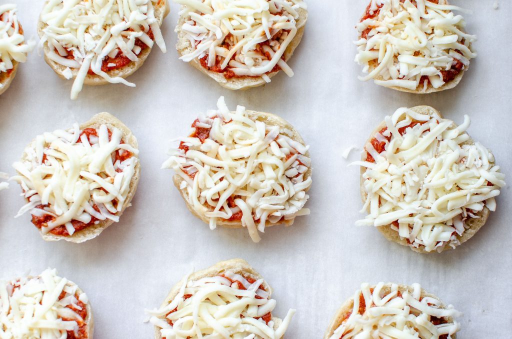 mini uncooked bagels bites with sauce and shredded mozzarella cheese on parchment paper