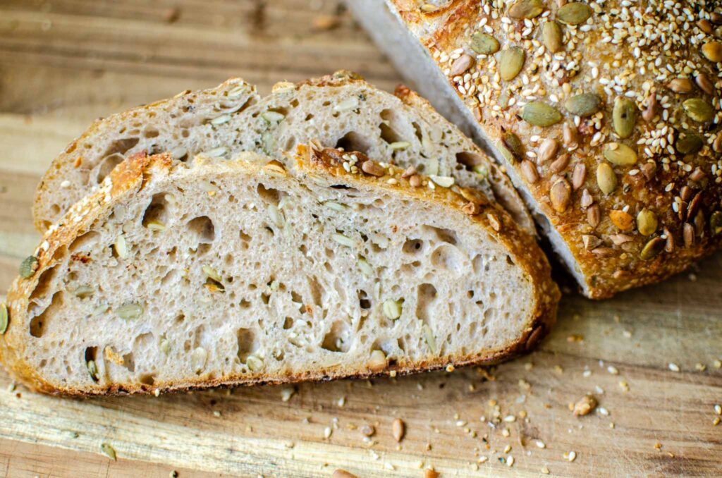 Slices of seeded multigrain sourdough bread on wood cutting board next to loaf