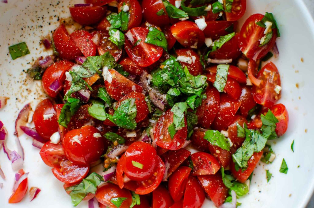 cut tomatoes, basil leaves and red onion mixed in a bowl with freshly crack black pepper and salt flakes on top