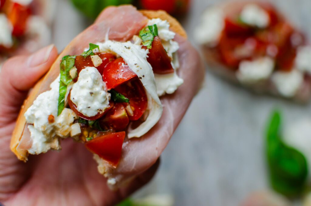 holding a piece of Easy Burrata Bruschetta and Prosciutto Toast with a bite taken