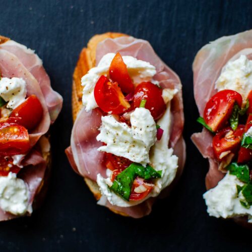 Three burrata bruschetta and prosciutto toasts on black slate.