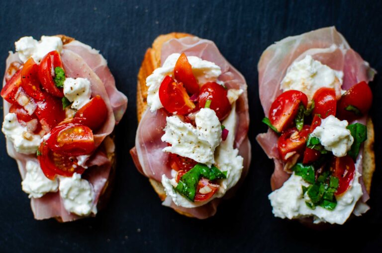 Three burrata bruschetta and prosciutto toasts on black slate.