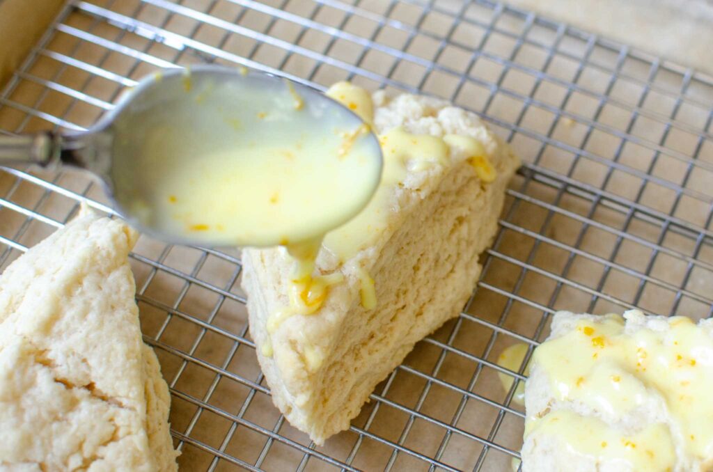 spooning the sweet orange glaze over a baked sourdough discard orange scone