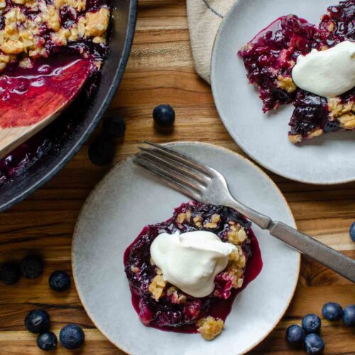 Two plates and a cast iron skillet with blueberry rhubarb crisp and whipped topping.