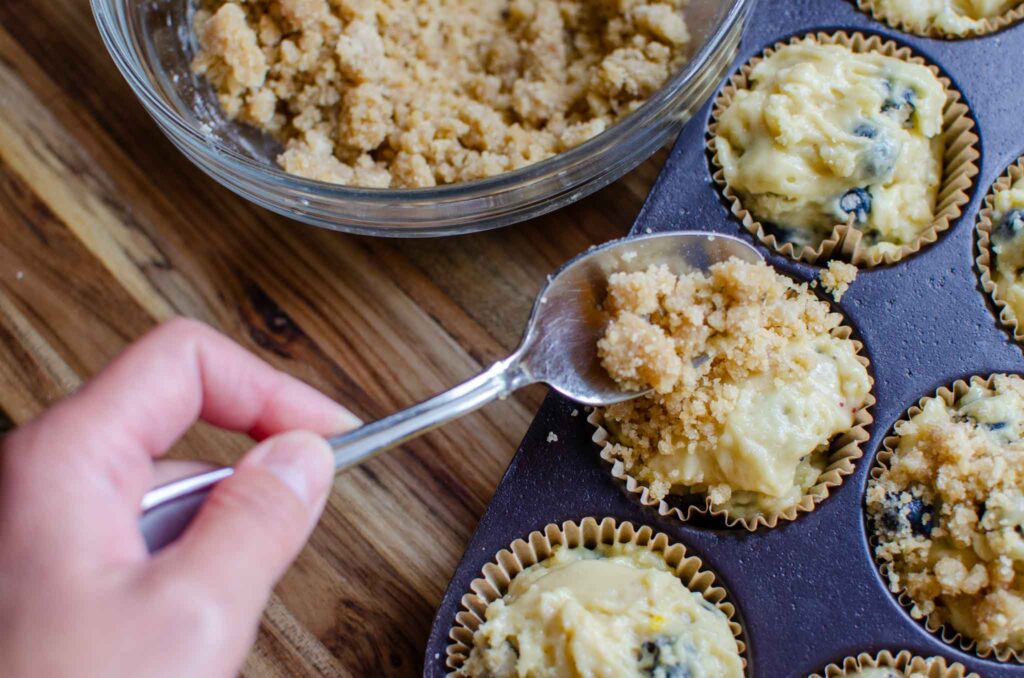 Spooning crumb mixture over the top of sourdough blueberry muffins in pan