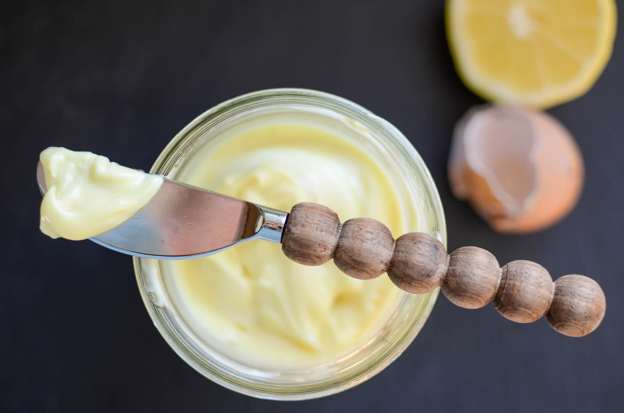 A mason jar of homemade mayo with spreading knife, egg shells and half of lemon