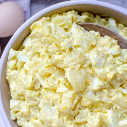 classic potato salad in a round clay pan with a wooden serving spoon. An egg and potatoes next to the serving dish.