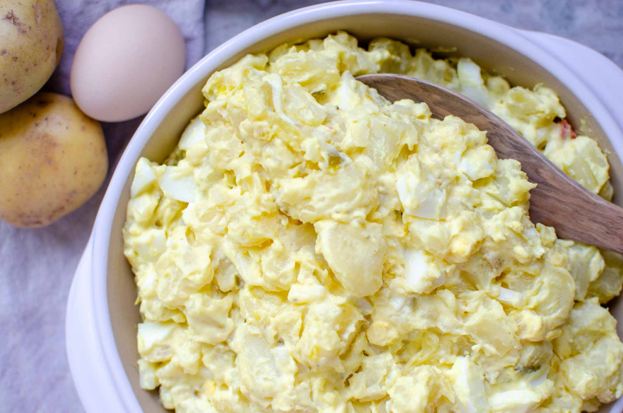 classic potato salad in a round clay pan with a wooden serving spoon. An egg and potatoes next to the serving dish.