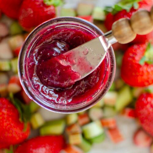 a jar of mixed berry rhubarb jam with spreader with fruit all around the jar