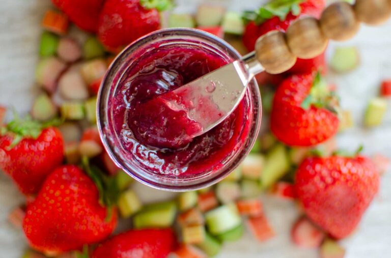 a jar of mixed berry rhubarb jam with spreader with fruit all around the jar