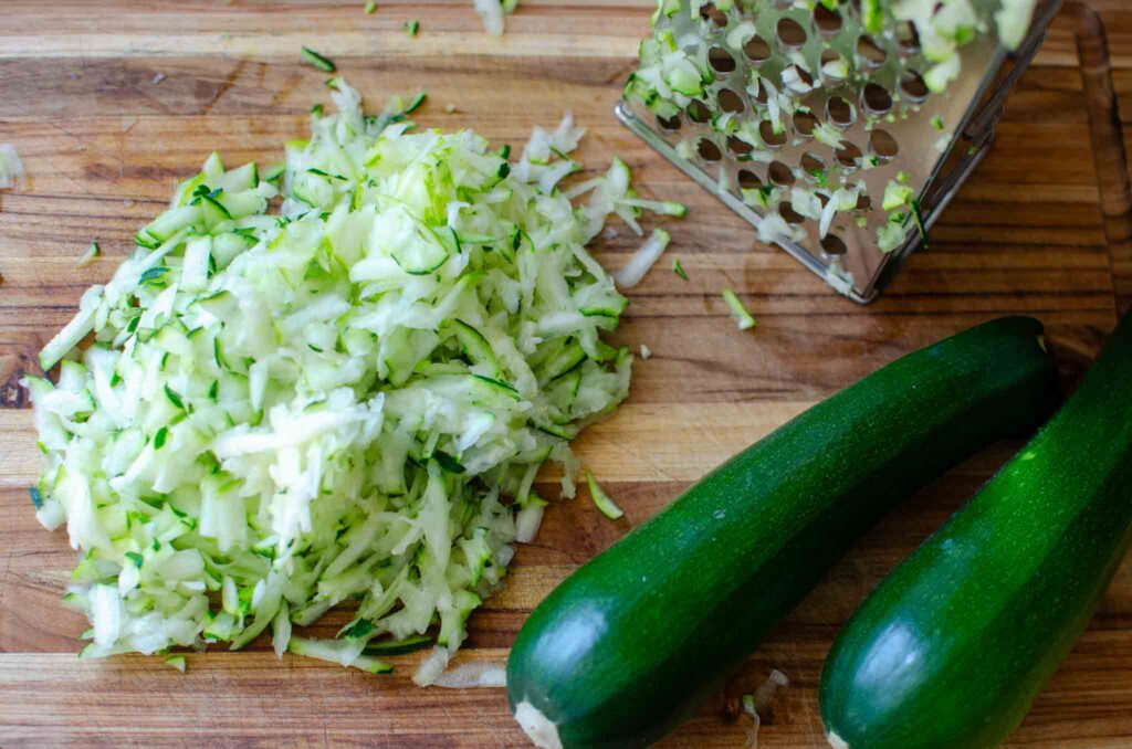 whole and grated zucchini and box grater on a wooden cutting board.