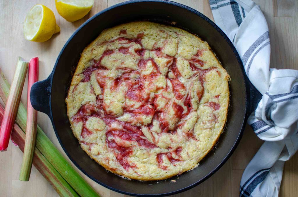 Baked rhubarb spoon cake in cast iron skillet with rhubarb stalks and cut lemon