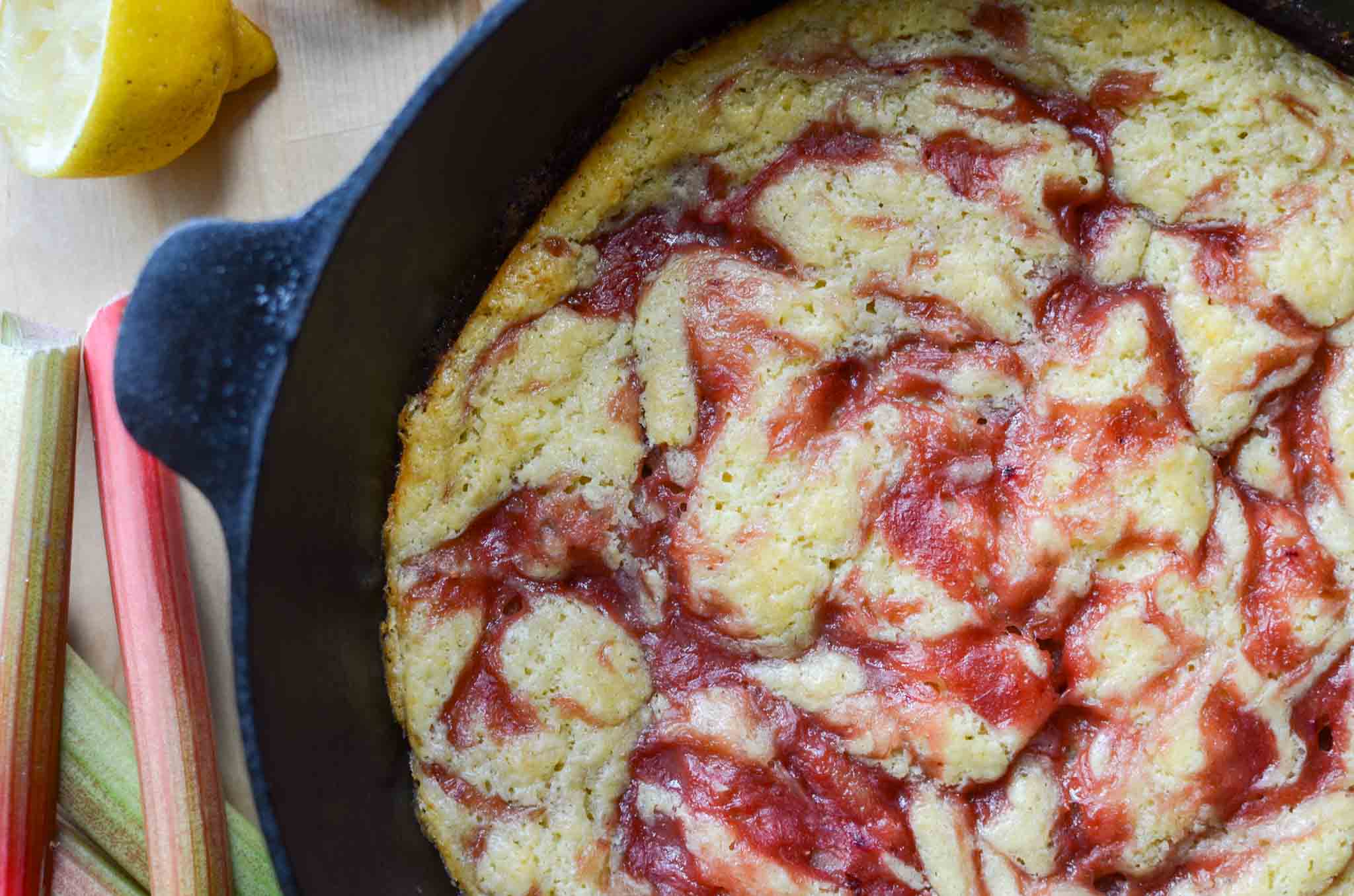 Rhubarb spoon cake in a cast iron skillet with rhubarb stalks and cut lemon