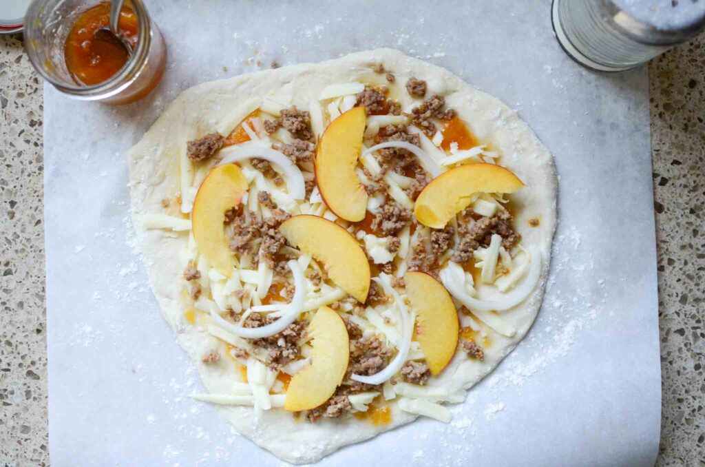 Italian sausage, onion, and peach slices on pizza dough on a piece of parchment paper