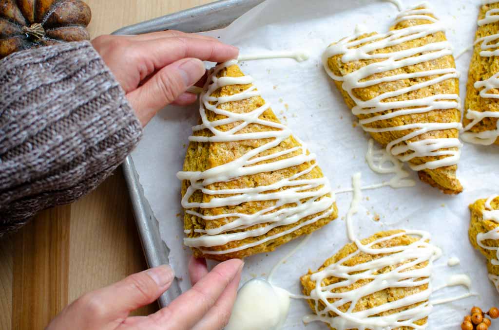 hands picking up a sourdough pumpkin scone with cream cheese glaze from a baking sheet.