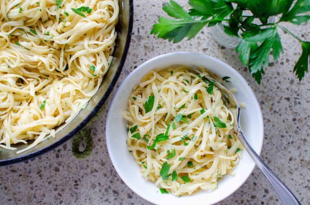 a white bowl of pasta aglio e olio next to a pan of the pasta