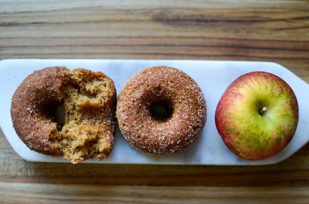 a broken apple cider donut on its side, a whole donut and an apple on a serving plate