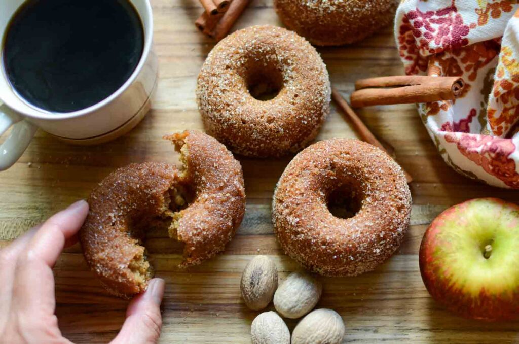 a hand grabbing a torn apart sourdough apple cider donut, with cinnamon sticks and a cup of coffee close by.