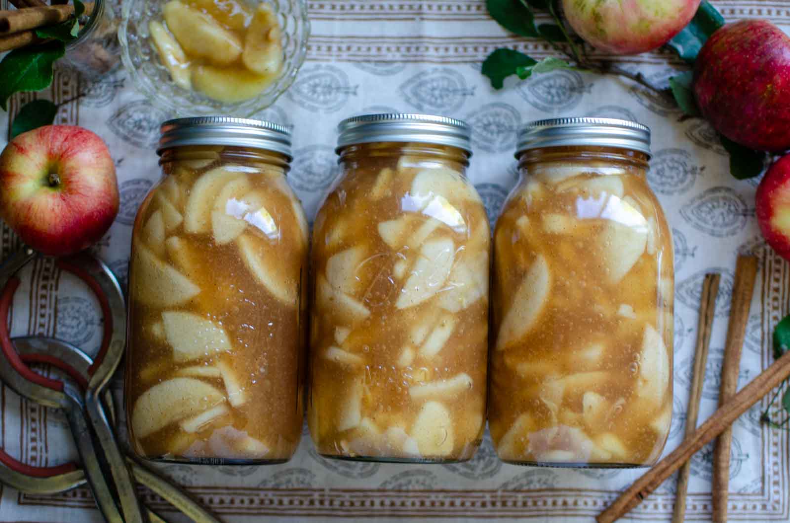 apple pie filling canned in glass mason jars with apples and cinnamon sticks next to them.