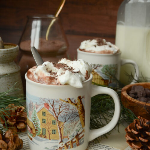 two mugs of hot cocoa with a bowl of chocolate chips and pine needles next to it