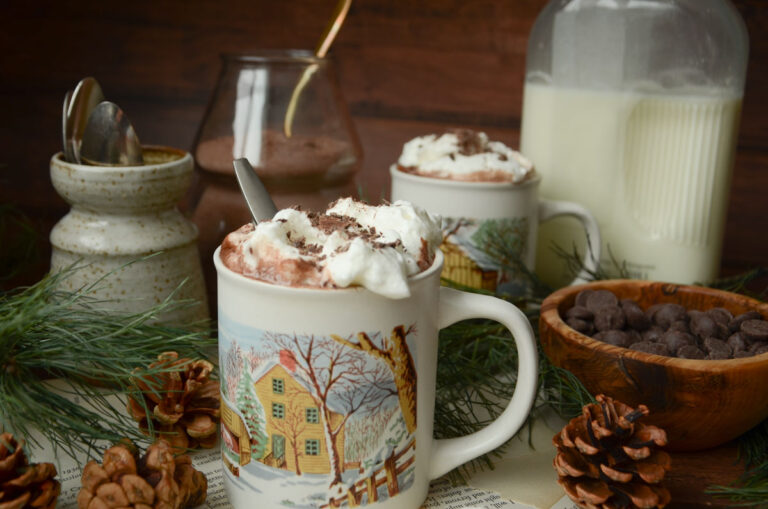 two mugs of hot cocoa with a bowl of chocolate chips and pine needles next to it