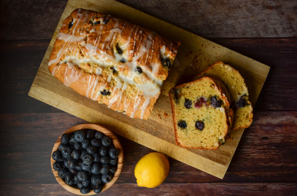 lemon blueberry sourdough bread loaf on a wood cutting board with bowl of blueberries and whole lemon