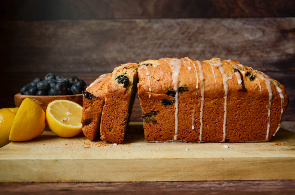 Sourdough lemon blueberry quick bread loaf on wood cutting board with cut lemons and a bowl of blueberries