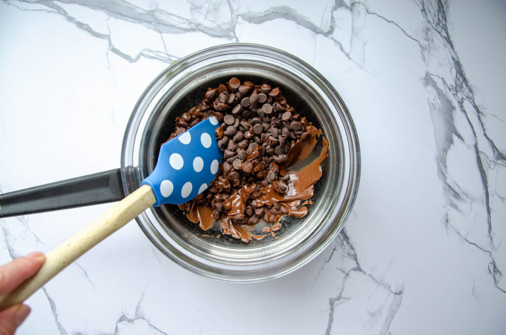 melting chocolate chips in a glass bowl set over a saucepan, mixing with a blue silicone spatula