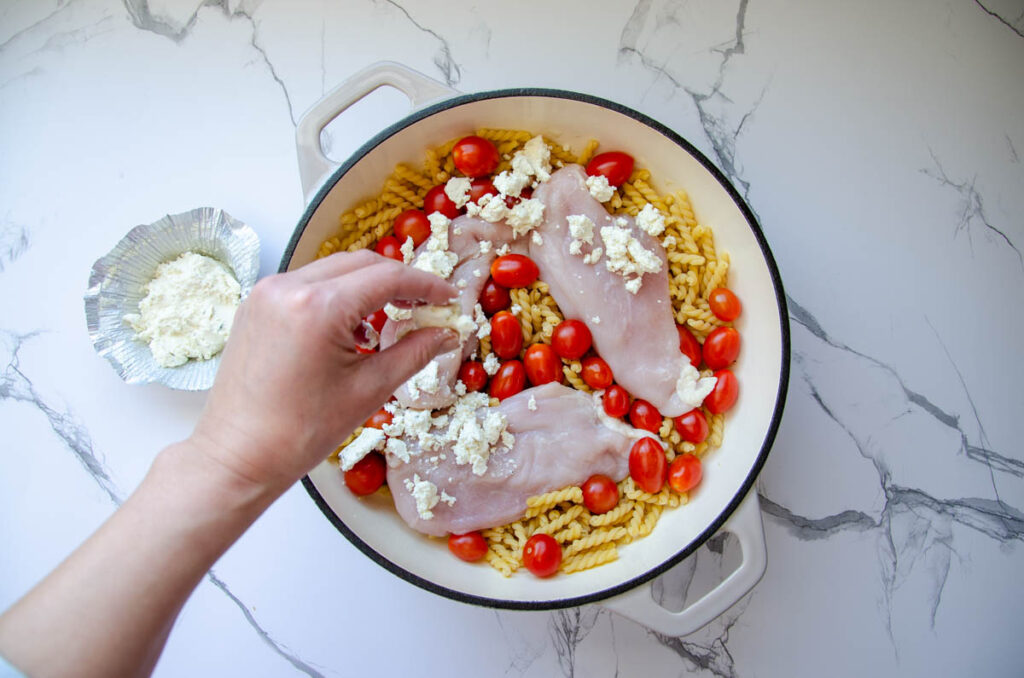 Crumbling Boursin cheese over chicken breasts, pasta, and tomatoes in enameled pan