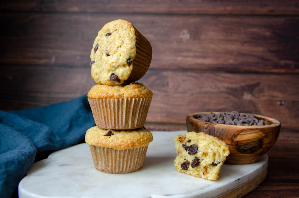 three sourdough chocolate chip muffins stacked on top of one another with a bowl of chocolate chips and half of a muffin next to it. 